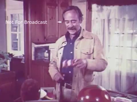The still from the 16mm film 'Toy Safety - Consumer Product Safety Commission PSA' from the 1970s features a man with a mustache, dressed in a light-colored jacket, standing in a cozy indoor setting, likely a living room or kitchen. He appears to be demonstrating a toy to a child, who is partially visible, sitting at a table cluttered with colorful items, including balloons. The environment suggests a familial or educational atmosphere focused on toy safety awareness.