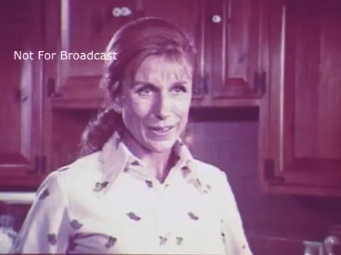 The still from the 16mm print of 'Toy Safety' features a woman with long hair and a patterned blouse, appearing to address the viewer with a concerned expression. The background suggests a kitchen setting, with wooden cabinetry visible. This PSA aims to raise awareness about the importance of toy safety, likely delivering key messages on how to ensure children's toys are safe for use. The overall tone is informative and cautionary, typical of public service announcements from the 1970s.