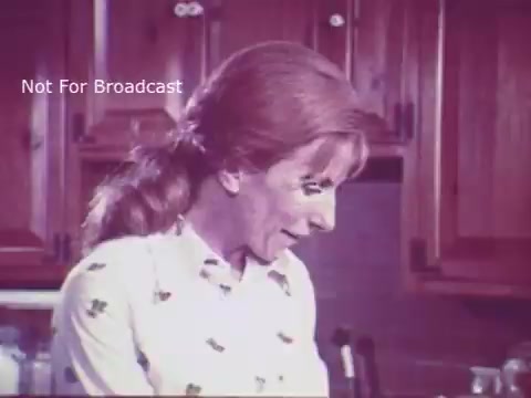 The still from the 16mm film 'Toy Safety - Consumer Product Safety Commission PSA - 1970's' features a woman in a kitchen setting, appearing engaged and attentive. She has long, wavy hair and is wearing a patterned blouse. The context emphasizes the importance of toy safety, likely discussing hazards and recommendations for parents to ensure child safety. The visual style reflects the aesthetic of the 1970s, with warm, muted colors typical of the era.