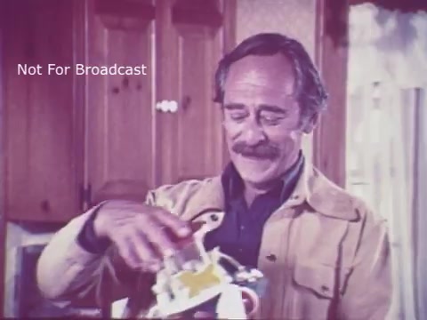 The still from the 1970s PSA 'Toy Safety' features a man in a kitchen setting, smiling as he interacts with a toy. The background suggests a typical home environment with wooden cabinetry. The man appears engaged, highlighting a focus on toy safety, likely addressing parents or caregivers about the importance of checking toys for safety hazards. The overall tone is educational and preventative.