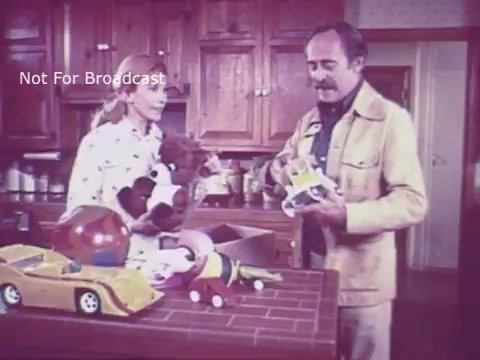The still from the 16mm print of the 'Toy Safety - Consumer Product Safety Commission PSA' shows a man and a woman in a kitchen environment, surrounded by various toys. The woman is holding a stuffed animal, while the man appears to be examining a toy vehicle. The scene emphasizes toy safety, likely discussing concerns and guidelines related to safe play for children. The warm lighting and homey setting reflect the educational tone of the PSA from the 1970s.