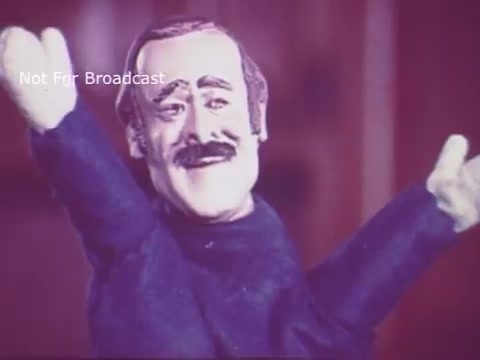 The still from 'Toy Safety - Consumer Product Safety Commission PSA - 1970's' features a character with a distinct mustache and an expressive face. The character appears to be animated or a puppet, demonstrating a friendly demeanor with arms raised. The colors in the image have a vintage quality typical of 16mm film, contributing to a nostalgic feel. The context suggests the character is likely conveying an important safety message regarding toys, aimed at educating viewers about consumer product safety.