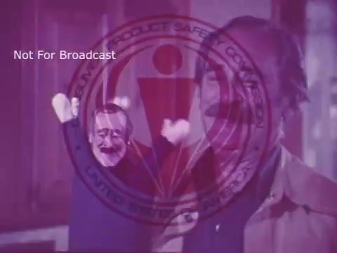 The still from the 16mm print of the PSA 'Toy Safety' features a man, seemingly engaged in a playful demonstration with a puppet, emphasizing the importance of toy safety. The Consumer Product Safety Commission logo is prominently overlaid, indicating that the message is intended for public awareness regarding safe toys for children. The overall tone suggests a light-hearted yet informative approach to educating viewers on potential hazards in toys.