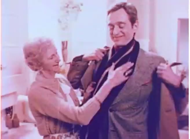 The still from the 1970s Alka-Seltzer commercial features a lighthearted scene in a cozy setting. An older woman is helping a well-dressed man with his jacket, showcasing a warm interaction between them. The color palette leans towards soft pastel tones, typical of that era, enhancing the nostalgic feel. This captures a homely and inviting atmosphere, fitting for a product pitched for comfort and relief. Follow us on Twitter @FromFilm for more vintage insights!