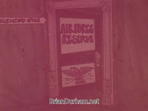 The still features a door with a sign that reads 'AIR FORCE RESERVE.' There’s an eagle symbol on a lower panel, suggesting military affiliation. To the left, an arrow points to 'Recruiting & Info,' indicating the purpose of the space. The overall look has a retro feel typical of 1960s advertising.