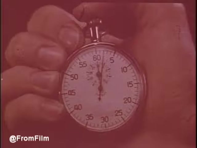 The still from the 'M*A*S*H Leukemia Public Service Announcement' features a close-up of a hand holding a stopwatch. The stopwatch is set to nearly one minute, emphasizing the urgency of the message. The color tone is warm, creating a reflective atmosphere. This image is likely symbolic, representing a countdown or race against time to find a cure for leukemia, echoing the sentiments expressed by Loretta Swit and Mike Farrell in the announcement.