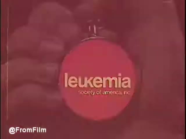 The still features a close-up of a circular clock or watch, prominently displaying the text 'leukemia' and 'society of america, inc.' against a red background. This image likely symbolizes urgency and hope in the context of the public service announcement, emphasizing the ongoing fight against leukemia and the efforts to find a cure. The overall tone is serious, reflecting the gravity of the cause.