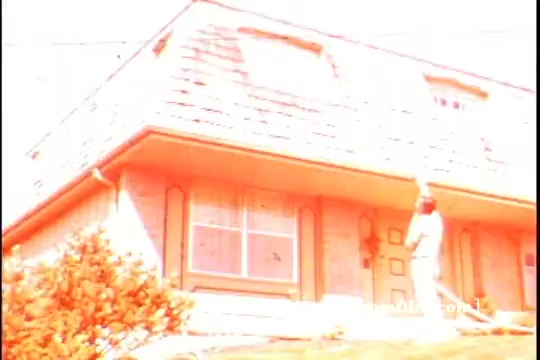 The still from the 16mm 'Jetx car washing commercial - 1970' features a brightly lit house with a sloped roof. A person, dressed in light clothing, is seen standing outside, possibly washing a car or engaging in outdoor maintenance. The overall color palette appears heavily saturated, giving the image an overexposed, artistic quality. The scene captures a suburban setting, typical of the era.