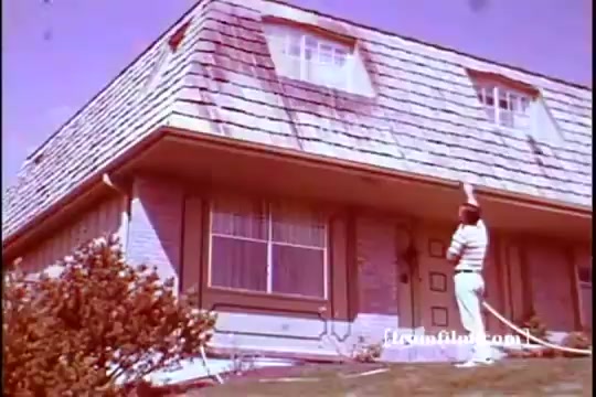 The still from the 1970 Jetx car washing commercial features a man in a striped outfit, standing in front of a two-story house with a sloped roof. He holds a hose, likely for washing, while looking up at the roof. The setting appears sunny, with greenery in the foreground and large windows on the house, suggesting a suburban neighborhood. The overall tone is vibrant, indicative of the era.