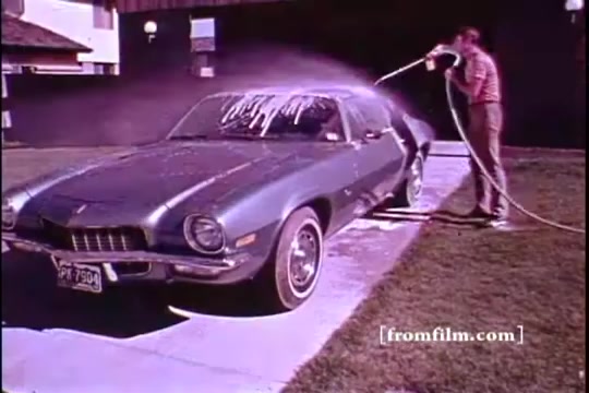 The still features a vintage green car being washed. A person is spraying foam and water onto the vehicle using a hose. The setting appears to be a driveway in front of a house, with a clear sky. The scene reflects the casual, suburban lifestyle typical of the 1970s. The image has a warm color tone, characteristic of 16mm film.