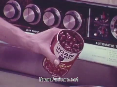 The still features a hand holding a can of 'Joan of Arc Kidney Beans.' The can is prominently displayed, showcasing its label. In the background, there are dials of an appliance, suggesting a kitchen setting. The overall tone reflects a vintage 1960s aesthetic, emphasizing home cooking.