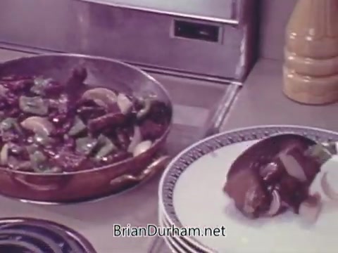 The still from 'Joan Of Arc Kidney Beans - 1960s' features a skillet filled with a savory dish, likely containing kidney beans, alongside chopped vegetables such as green peppers and onions. A serving is placed on a plate, suggesting a focus on home cooking and meal presentation typical of that era. The kitchen setting is simple and functional, indicative of the 1960s style.