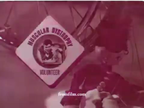 The still from the PSA 'Muscular Dystrophy: No Strings' features a prominent diamond-shaped badge with the text 'MUSCULAR DYSTROPHY' at the top and 'VOLUNTEER' at the bottom. The badge includes an image likely depicting a figure related to the muscular dystrophy cause. The overall color tone is reddish, suggesting a vintage feel, typical of early 1970s public service announcements. The faded quality indicates age, common in archival film footage. The context emphasizes the call to action for viewers to support the cause by getting involved with volunteer efforts, particularly associated with Jerry Lewis.