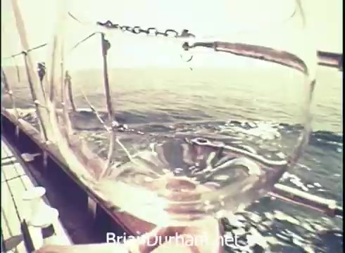 The still from the 1971 Lancer Wine commercial features a close-up shot of a glass of wine, with a blurred view of ocean waves and a boat in the background. The transparent glass highlights the wine's hues while capturing the nautical setting, suggesting enjoyment of the drink amidst a scenic maritime environment. The composition emphasizes both the wine and the tranquil sea.