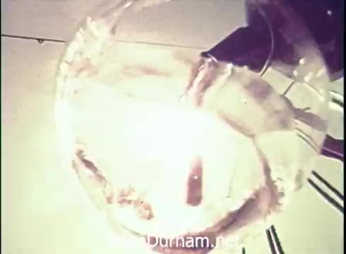 The still features a close-up shot of a wine glass being filled, with a stream of liquid pouring from a bottle. The glass reflects light, creating a sparkling effect, suggesting freshness and allure. The background is minimal, focusing attention on the wine being served, indicative of a classic advertising style typical of the early 1970s.