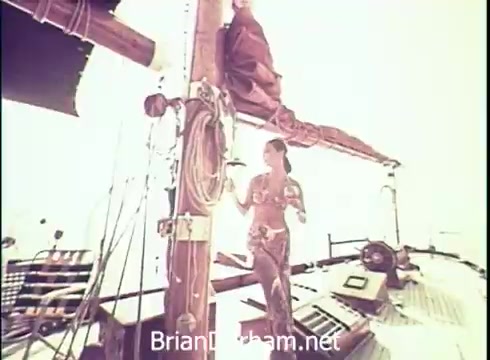 The 16mm still from the 1971 Lancer Wine commercial features a woman on a sailboat. She is dressed in vibrant, tropical attire and appears to be enjoying her surroundings. The bright, sunlit scene emphasizes a carefree, leisurely atmosphere, aligning with the branding of Lancer Wine. The boat's rigging and deck details add to the nautical theme of the advertisement.