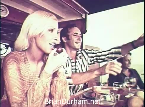 The still from the 1971 Lancer Wine commercial features a relaxed gathering where a blonde woman bites into an apple while a man in a striped shirt joyfully points, likely engaged in conversation. They are seated around a table adorned with glasses of wine, suggesting a casual, social atmosphere aboard a boat or similar setting. The overall mood is cheerful and carefree, characteristic of advertising from that era.