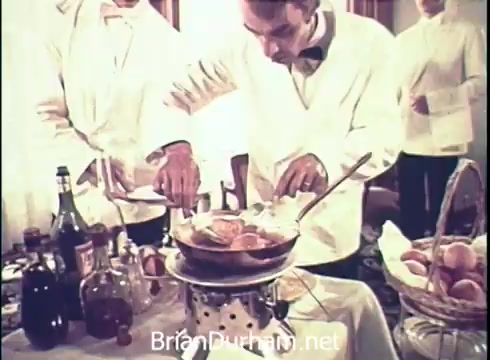 The 16mm still from the 1971 Lancer Wine commercial features a well-dressed man in a tuxedo preparing food in a stylish dining setting. He is focused on a pot, likely containing a dish, while surrounded by bottles of wine and a basket of eggs. The atmosphere suggests an upscale dining experience, emphasizing the pairing of wine with gourmet cuisine.
