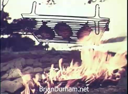 The still from the 'Lancer Wine Commercial - 1971' features a grill suspended over an open fire. The grill holds three rounded items, likely food, and flames rise from below, creating a vibrant and inviting atmosphere. The background appears simplified, emphasizing the grilling scene, which suggests a focus on outdoor enjoyment and pairing with Lancer wine.