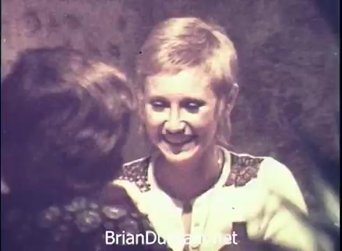 The still from the 'Lancer Wine Commercial - 1971' features a young woman with short hair smiling warmly at another person. The setting has a vintage look, typical of the 1970s. The background appears slightly blurred, focusing attention on the interaction between the two individuals. The overall vibe suggests a friendly and inviting atmosphere, likely intended to promote the wine brand's social appeal.