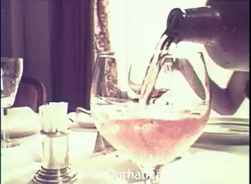 The still features a close-up of a glass being filled with a light pink liquid, likely wine, from a bottle. The table is set with additional glasses and a salt shaker in the foreground. The background suggests an elegant dining setting, with soft lighting creating a warm atmosphere. The scene conveys a sense of relaxation and enjoyment, characteristic of a commercial for Lancer Wine from 1971.