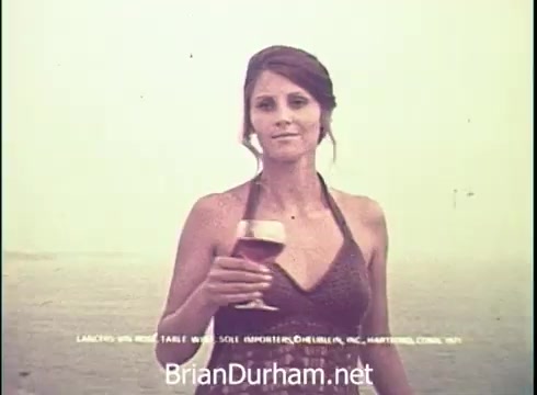The still from the 1971 Lancer Wine commercial features a woman holding a glass of wine. She has long hair styled elegantly and is wearing a halter top that suggests a casual yet sophisticated setting. The background is hazy, possibly suggesting a seaside or outdoor atmosphere, enhancing the relaxed vibe of the advertisement. The text at the bottom indicates it promotes Lancer's wine, emphasizing its appeal for table and casual drinking.