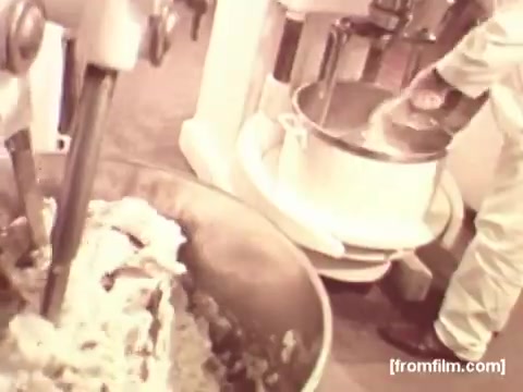 The still from the Pepperidge Farms Apollo 17 commercial features industrial kitchen equipment in action, focusing on the mixing process. The foreground shows a large bowl of dough being mixed, while a second mixing bowl is visible in the background, suggesting a busy baking environment. The overall tone of the footage is warm and nostalgic, reflecting an emphasis on artisanal bread-making. The lack of voiceover emphasizes the visual aspects of the production process.