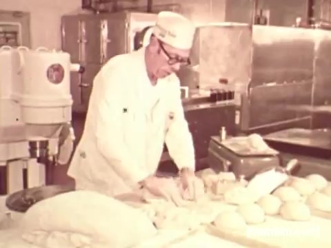 In the still from the 'Pepperidge Farms - Apollo 17' commercial, a baker in a white uniform and cap is seen working in a kitchen. He is focused on shaping dough on a countertop, surrounded by several loaves of bread. The kitchen appears well-equipped, with shiny metallic surfaces and baking tools. The scene emphasizes the craftsmanship and care involved in bread-making, in line with the brand's legacy.