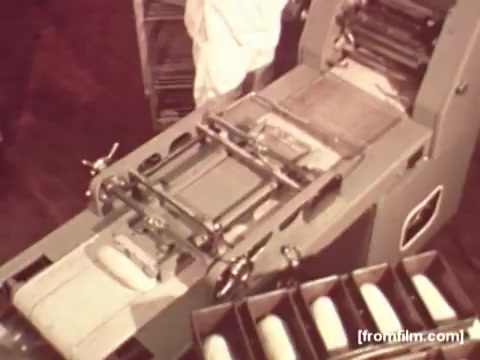 The still from the 'Pepperidge Farms - Apollo 17' commercial features a top-down view of a bread production line. An industrial machine, equipped with rollers and trays, processes loaves of bread as they move through the assembly. The scene is captured in a nostalgic, muted color typical of 16mm film, emphasizing the craftsmanship involved in bread making. The absence of voiceover suggests a focus on the visual narrative, highlighting the industrial scale and precision of the production process.