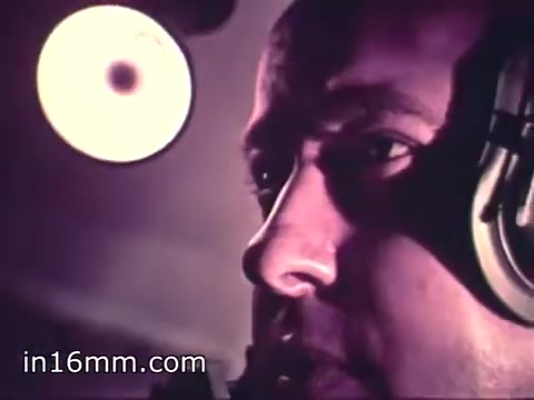 The still from 'Air Force Reserve Recruiting - 1968 - Air Crews' features a close-up of a male individual, likely a pilot or crew member, wearing headphones. His face is illuminated by a nearby light, emphasizing his serious expression and focused demeanor. This suggests a moment of concentration, possibly while communicating or operating equipment. The warm color tones and vintage film quality contribute to the documentary's historical context, highlighting the importance of air crew roles in the Air Force Reserve during that era.