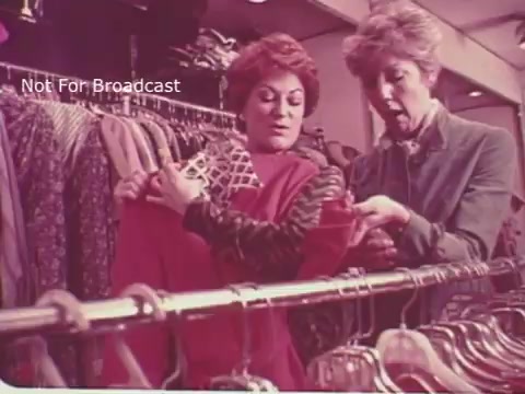 The still features two women in a clothing store, examining garments. One woman appears enthusiastic, holding up a colorful outfit, while the other looks surprised or concerned, pointing at an item. The context of the PSA emphasizes the importance of reading medicine labels correctly to ensure proper use, akin to carefully choosing the right clothing.