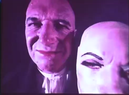 The still features a close-up of a man with a sinister smile alongside a wax figure. The man's expression is unsettling, contrasting with the lifeless face of the wax figure, which has exaggerated features. The colors are muted, typical of a 16mm print, adding to the eerie atmosphere of the scene, suggesting themes of horror and deception.