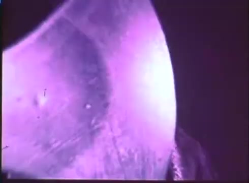 The still features a close-up of a metallic surface, likely a prop or set piece, displaying an iridescent sheen with purple hues. The lighting creates dramatic shadows and highlights, contributing to a mysterious and eerie atmosphere typical of the horror genre. The texture suggests wear, adding to the vintage quality of the 16mm print from the trailer of 'Terror in the Wax Museum.'