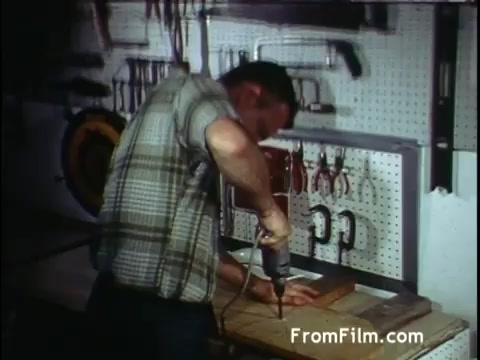 The still from the 'C and P Telephone' commercial features a man in a plaid shirt working in a workshop. He is using a power drill on a piece of wood resting on a bench, surrounded by various tools hanging on a pegboard. The setting suggests a focus on home improvement or craftsmanship, aligning with the telecommunications theme of connecting people and services. The visual style is indicative of the early 1970s era, emphasizing practicality and hands-on work.