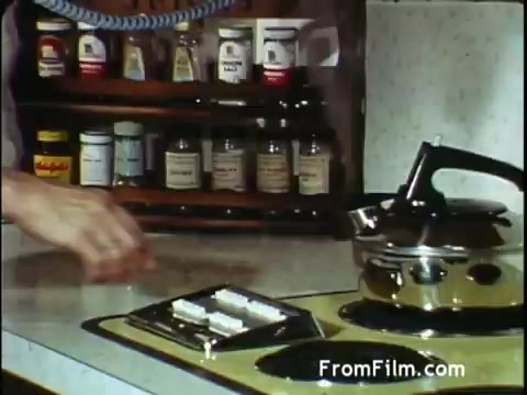 The still from the C&P Telephone commercial features a close-up of a kitchen countertop. A hand is seen reaching for a small appliance, presumably a stove, while a kettle is visible beside it. In the background, a shelf filled with various spice containers adds a homely touch to the setting. The scene conveys a domestic and everyday life atmosphere, typical of advertisements from the early 1970s, emphasizing convenience and family-friendly themes.