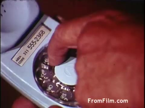The still from the C&P Telephone commercial features a close-up of a person’s hand dialing a rotary telephone. The phone has a classic design typical of the early 1970s, with a visible dial and a label indicating a phone number. This commercial likely aimed to showcase the ease of using their telephone service during a time when rotary phones were common. The overall mood reflects a nostalgic connection to communication from that era.