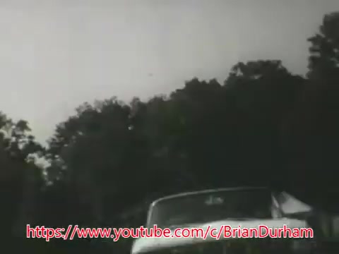 The still from 'Underwater Car PSA - 1969' features a classic automobile parked near a body of water, with trees in the background. The setting appears somber and serious, reflecting the grave topic of the PSA. The voice-over advises viewers on how to react if their car submerges, emphasizing the importance of not panicking and exiting through the windows. The visual and auditory elements combine to convey a crucial safety message regarding drowning in submerged vehicles.