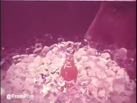 The still from the 1970s Orange Crush TV commercial features a bottle of Orange Crush nestled in a bed of ice cubes, surrounded by a warm, soft hue. The image evokes a refreshing, vibrant essence, characteristic of nostalgic advertising. The composition focuses on the bottle, emphasizing its appeal as a cool drink. The texture of the ice contrasts with the smooth glass, enhancing the visual invitation to enjoy the beverage. Follow us on Twitter @FromFilm.