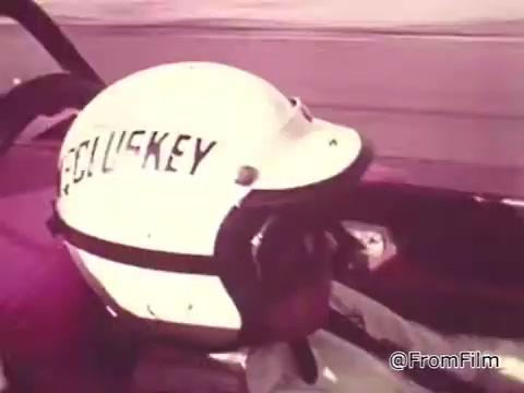 The still shows a close-up of a race car driver, Roger McClusky, wearing a white helmet with his name emblazoned on the side. He appears focused and determined, likely in an action-packed moment from the commercial promoting Chase and Sandborn Coffee. The background suggests high-speed racing, characteristic of the 1960s motorsport scene. The print quality reflects the vintage 16mm format, adding a nostalgic feel to the image.