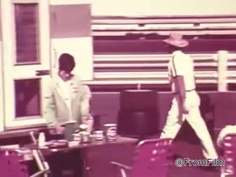 The still captures a scene from a 1960s Chase and Sandborn Coffee commercial featuring Roger McClusky, a race car driver. It shows two figures in a setting suggestive of a racetrack or pit area. One person, dressed in a light-colored suit, appears to be organizing items on a table, while another figure, wearing a straw hat and light clothing, walks past. The color tones are characteristic of the era, and the overall vibe is relaxed but focused on coffee, linking it to the racing theme.