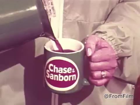 The still captures a scene from a 1960s Chase and Sanborn Coffee commercial featuring race car driver Roger McClusky. It shows a hand pouring coffee from a green coffee pot into a matching green mug emblazoned with the Chase and Sanborn logo. The setting suggests a casual, automotive vibe, reflective of the era's advertising style.
