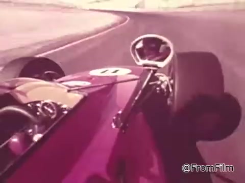 The still features a close-up view from the cockpit of a race car, showcasing the steering wheel and dashboard. The vibrant colors emphasize the sleek design of the car, which is likely a racing model from the 1960s. The background suggests a racetrack, enhancing the thrilling atmosphere. This image captures the high-speed excitement associated with the Chase and Sandborn Coffee commercial featuring race car driver Roger McClusky.