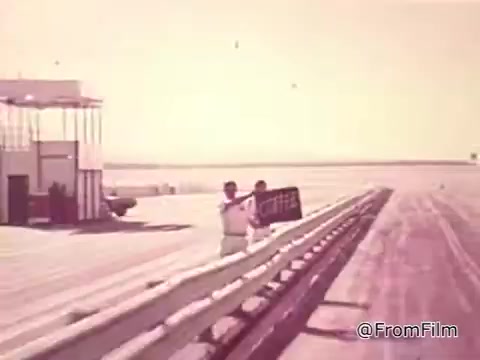 The still features a scene from a 1960s Chase and Sandborn Coffee commercial, showcasing a race track environment. A racetrack worker is seen waving a sign, likely indicating the beginning or end of a race event. The background is minimalist, emphasizing the emptiness of the track and the influence of the coffee brand, with a feeling of nostalgia characteristic of the era. The colors are muted, typical of 16mm film prints from that time.