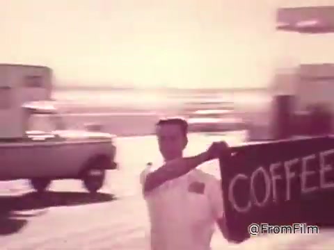 The still from the 1960s Chase and Sandborn Coffee commercial features a man in a white shirt, energetically holding up a sign that reads 'COFFEE.' The scene is set in a lively environment reminiscent of a racetrack, likely in Las Vegas, with vehicles in the background, capturing the dynamic atmosphere of the era. The image, taken from a 16mm print, showcases the vintage style and promotional flair typical of commercials from that time.