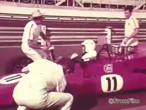 The still captures a scene from a 1960s Chase and Sanborn Coffee commercial featuring race car driver Roger McClusky. It shows a red race car with the number 11 as crew members in white uniforms assist the driver, who is wearing a helmet. The backdrop suggests a racetrack, emphasizing the high-paced world of auto racing while promoting the coffee brand. The vintage quality of the 16mm print adds to the nostalgic feel of the era.