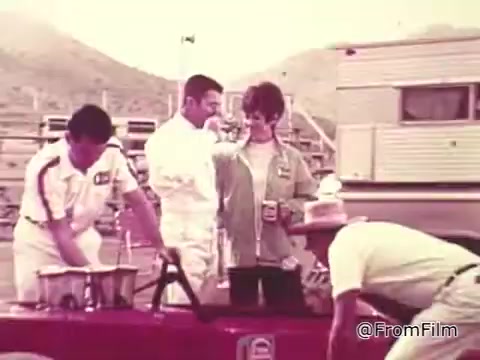 The still from the 1960s Chase and Sanborn Coffee commercial features a scene at a racetrack. Roger McClusky, a race car driver, is depicted interacting with a woman and two men in racing attire. They seem to be in a lighthearted moment, possibly enjoying coffee together. The backdrop includes a camper and mountains, typical of the 1960s racing atmosphere. Overall, it captures a blend of automotive culture and casual social interaction.