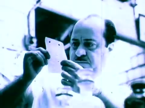 The still from the 'Scripto The Reading Pen Commercial' features a man intently examining a small card or piece of paper. He appears focused and possibly puzzled, indicating the importance of reading or understanding the text. The lighting is bright, giving the scene a slightly washed-out look, which adds an element of retro aesthetics typical of 16mm film. The setting suggests a workspace or industrial environment.