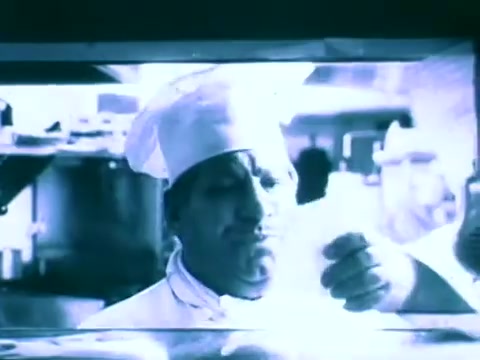 The still shows a chef in a kitchen, wearing a traditional white hat and jacket. He appears to be examining a piece of paper with a thoughtful or concerned expression. The image has a blue tint, contributing to a somewhat nostalgic or retro feel, characteristic of 16mm film. The kitchen background suggests a culinary context, likely showcasing a scenario related to reading or understanding recipes, tying into the theme of the Scripto Reading Pen.
