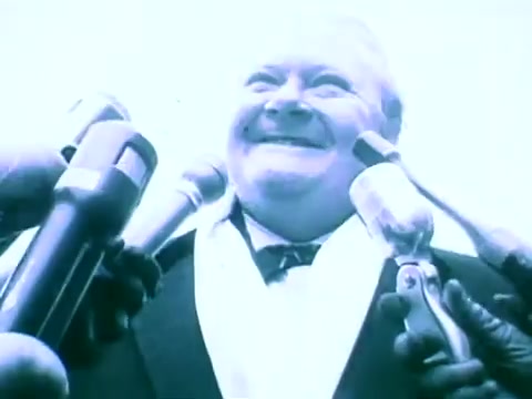 The still features a smiling man in a suit, surrounded by several microphones. The overall tone is bright and possibly exaggerated, suggesting a moment of public speaking or announcement. The context is likely promotional, highlighting the Scripto Reading Pen in a dynamic manner.