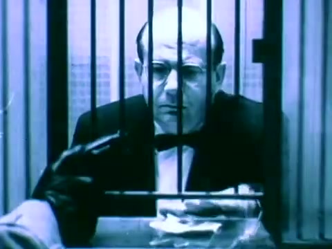 The still from 'Scripto The Reading Pen Commercial' depicts a man in a tuxedo sitting behind bars, conveying a sense of confinement or interrogation. The scene is filmed in a stark, blue-toned color palette, heightening the dramatic effect. A gloved hand is positioned in the foreground, possibly holding an item toward the man, suggesting tension or a pivotal moment in the commercial's narrative.